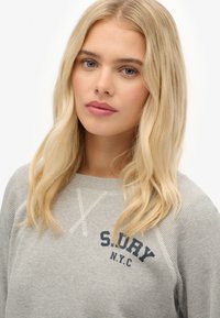 Suéter gris de punto acanalado, con texto en navy "S. L. U. R. Y. N.Y.C". Tiene cuello redondo y mangas raglán.