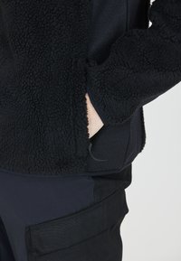 Giacca in pile nera con una superficie texturizzata e fluffy, dotata di una tasca laterale e un design a pannello liscio nella parte inferiore. Pantaloni scuri con tasche.