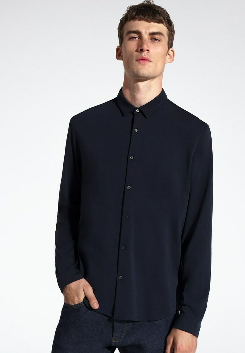 Camisa de botón azul marino oscuro con cuello, mangas largas y una textura suave; presenta una fila de botones y un corte relajado.