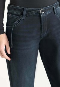 Jeans en denim bleu foncé avec une coupe slim, dotés d'une ceinture en tissu, de poches avant et d'une texture lisse. Détails de décoloration subtile et de couture.