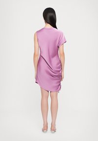 Robe en satin lavande à coupe décontractée, manches courtes, côtés froncés et ourlet arrondi, mettant en valeur les jambes. Des chaussures à talons hauts complètent le look.