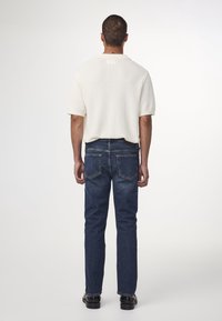 Hombre wearing camiseta de punto de manga corta blanca, jeans azules y zapatos negros, de pie con la espalda hacia la cámara contra un fondo blanco.