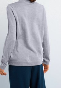 Pull en gris avec un col montant, des manches longues et des poignets côtelés. Fabriqué en matériau doux, présenté avec un pantalon bleu marine sur un fond clair.