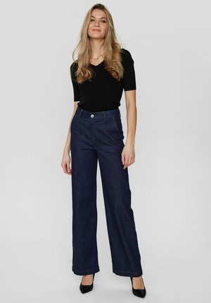 Sötétkék, széles szárú farmernadrág magas derekú fazonnal, gombos zárással és varrott részletekkel. Fekete, testhezálló toppal párosítva.