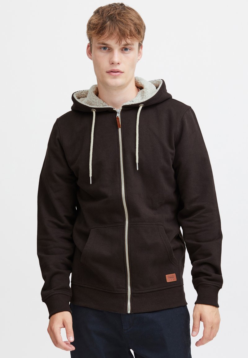 Bruine rits-hoodie met een fleece-gevoerde capuchon, trekkoordaccenten en zakken aan de voorkant. Bevat een klein merklabel aan de onderzijde.