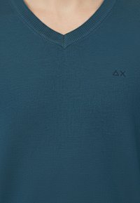 Maglione lavorato a maglia color teal con scollo a V, rifinito con bordi a coste e un piccolo logo in una tonalità più scura. Texture morbida, spessore medio.