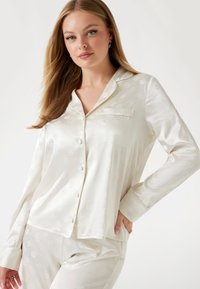 Weißes Satin-Pyjama-Set mit einem knöpfbaren Oberteil, das eine Tasche und ein dezentes Blumendesign aufweist, langen Ärmeln und passenden Hosen mit elastischem Bund.