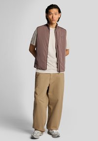 Leichte, ärmellose Weste in gedämpftem Mauve mit hochgeschlossenem Kragen und Seitentaschen, kombiniert mit einem hellen T-Shirt und beigen weiten Hosen.