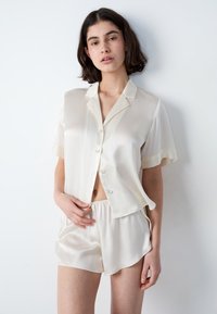 Kobieta z krótkimi ciemnymi włosami, ubrana w kremową satynową koszulę z krótkim rękawem zapinaną na guziki oraz pasujące szorty, stojąca na tle gładkiej białej ściany.