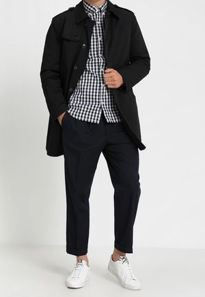 Homme portant un trench noir par-dessus une chemise à carreaux noir et blanc, un pantalon noir et des baskets blanches, debout avec une main dans la poche.