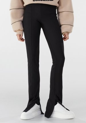 Person trägt schwarze, hoch taillierte Hosen mit vorderen Schlitzen über weißen Plateausneakern und einem beigen, übergroßen Sweatshirt.