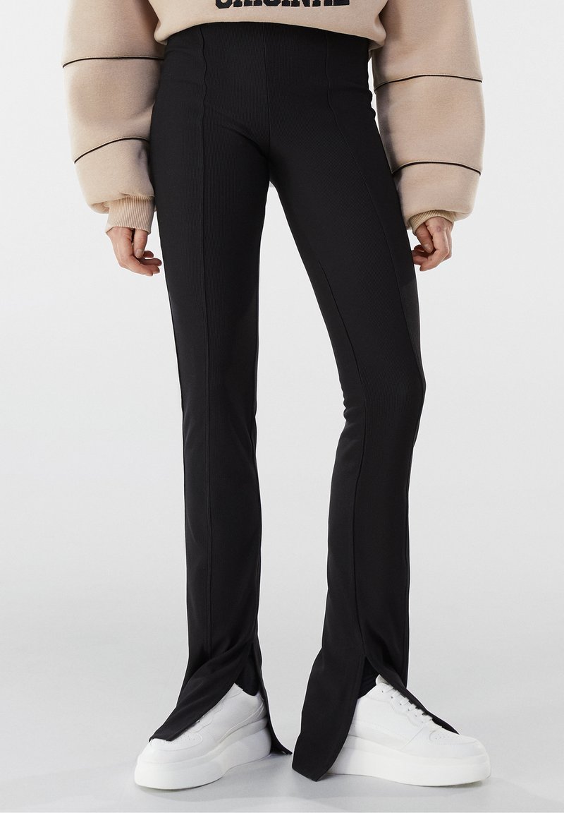 Personne portant un pantalon noir taille haute avec des fentes à l'avant, des baskets blanches à plateforme et un sweat-shirt beige oversize.