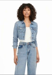 Giacca cropped in denim chiaro con due tasche frontali e chiusura a bottoni. Indossata sopra un top bianco, abbinata a jeans a vita alta.