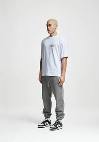 Tricou alb din bumbac cu un grafic mic pe piept, asociat cu pantaloni de trening gri și adidași negri cu alb. Aspect simplu și lejer.