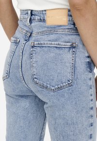 Primer plano de unos vaqueros de mezclilla azul claro con bolsillos traseros y una etiqueta de piel marrón en la cintura, combinados con una blusa blanca de canalé.