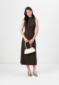 Brun ärmlös klänning med en åtsittande bodice och ett flödande kjol. Modellen håller en vit handväska med rundade kanter och bär beige spetsiga klackar.