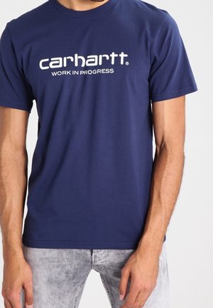 Man wearing a navy blue t-shirt with "carhartt WORK IN PROGRESS" printed in white on the chest and light gray jeans.