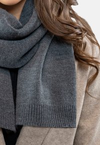 Sciarpa grigia lavorata a maglia con una texture liscia e bordi a costine, avvolta attorno al collo, abbinata a un cappotto beige.
