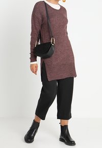 Maglione oversize bordeaux con fessure laterali, abbinato a pantaloni neri cropped larghi e stivali neri alla caviglia. La borsa è una piccola tracolla nera.