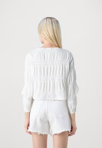 Blusa bianca corta con texture plissettata e accenti floreali ricamati sulle maniche e sull'orlo, abbinata a pantaloni corti in denim bianchi.