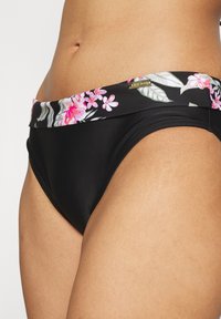 Μαύρο σλιπ μπικίνι με φαρδύ floral λάστιχο που διαθέτει ροζ και πράσινα σχέδια, κατασκευασμένο από απαλό, ελαστικό υλικό. Ορατή χρυσή λογότυπη λεπτομέρεια.