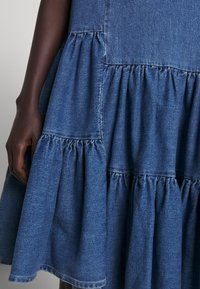 Džínové šaty s vrstevnatým volánkovým designem, ve středně modré barvě, s kontrastním šitím a jemnou texturou. Záběr zblízka na ruku, která drží látku.