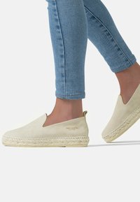 Beige slip-on espadrilles with a smooth suede finish, woven jute sole, and gold "FRED MARTIN" logo on the side. Texture and color contrast evident.