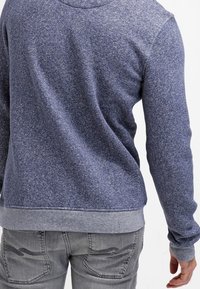 Blå mönstrad sweatshirt med en texturerad yta. Har en grå ribbad nederkant och ärmslut, samt en huva. Bärs med grå jeans.