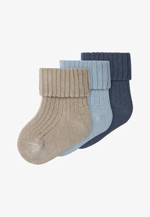 Trys sulankstyti riboto mezgimo kojinės smėlio, šviesiai mėlynos ir tamsiai mėlynos spalvų, išdėstytos kraštu persidengiantios ant balto fono.