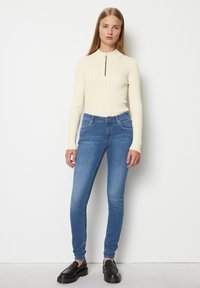 Tricou lung cu mânecă, cu dungi, de culoare crem, cu guler cu fermoar în jumătate, purtat cu blugi ajustați albaștri și loaferi negri cu talpă înaltă. Design de ținută simplu și modern.