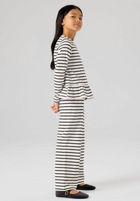 Fille debout de profil portant un haut à manches longues à rayures horizontales noires et blanches avec un ourlet à volants, ainsi qu'un pantalon large assorti et des chaussures noires.