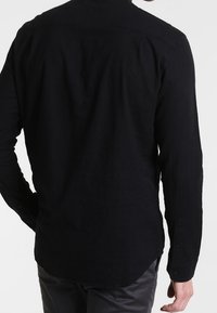 Maglietta nera a maniche lunghe con un tessuto texture, polsini con bottoni e design a orlo curvo. Cuciture visibili lungo le spalle e la schiena.