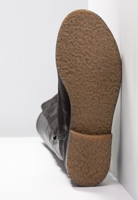 Botín de cuero negro con suela de goma texturizada, ligeramente inclinada. La parte inferior presenta patrones de agarre suave y letras de marca en relieve.