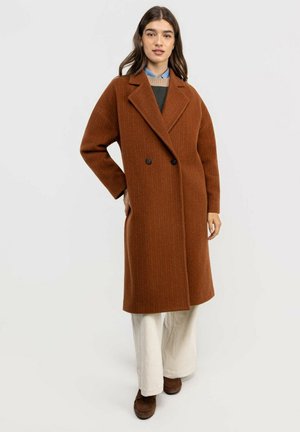 Mujer con un largo abrigo marrón cruzado sobre una blusa de capas y pantalones blancos de pierna ancha, de pie con una mano en la cadera.