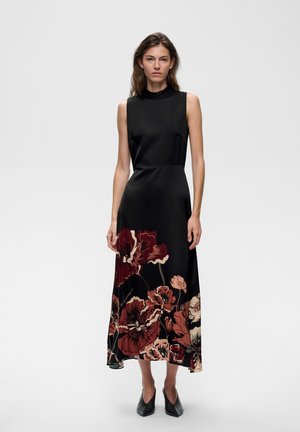Femme portant une robe midi noire sans manches avec un grand imprimé floral rouge et beige sur la jupe, debout devant un fond uni.
