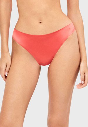 Femme portant un bas de bikini couleur corail, debout avec les bras détendus sur un fond uni gris clair.