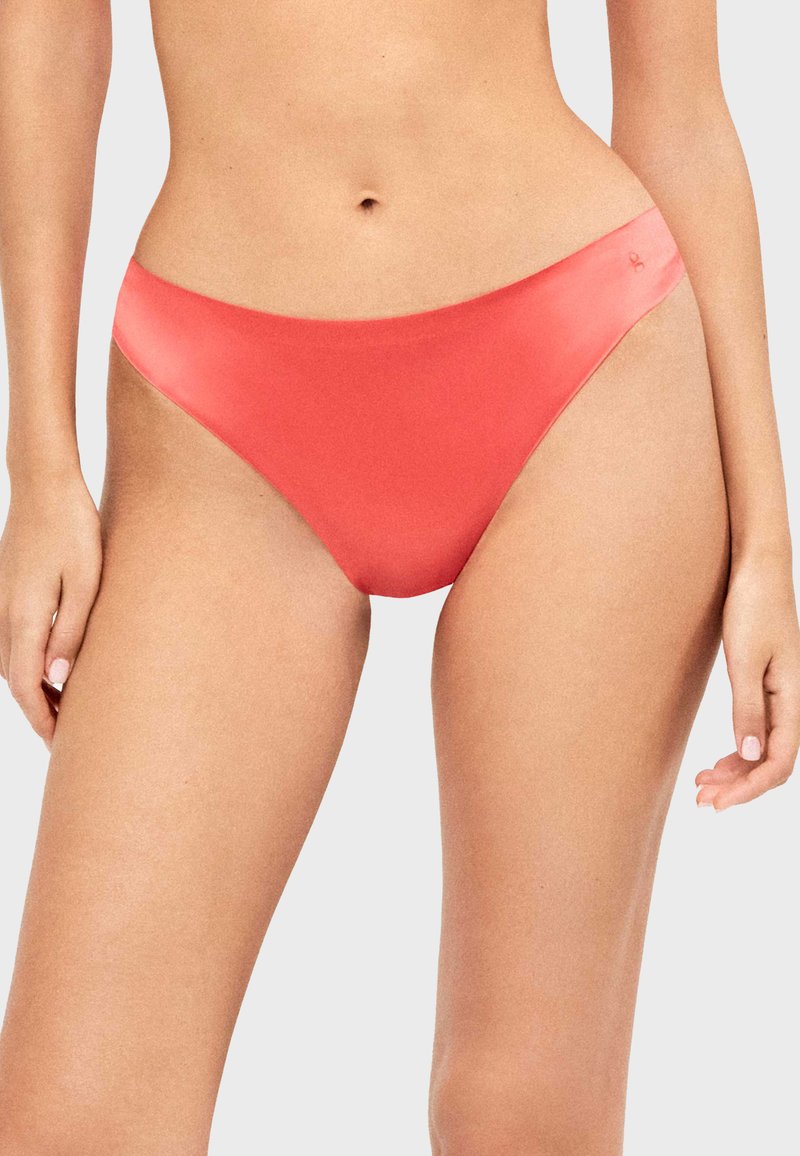 Femme portant un bas de bikini couleur corail, debout avec les bras détendus sur un fond uni gris clair.