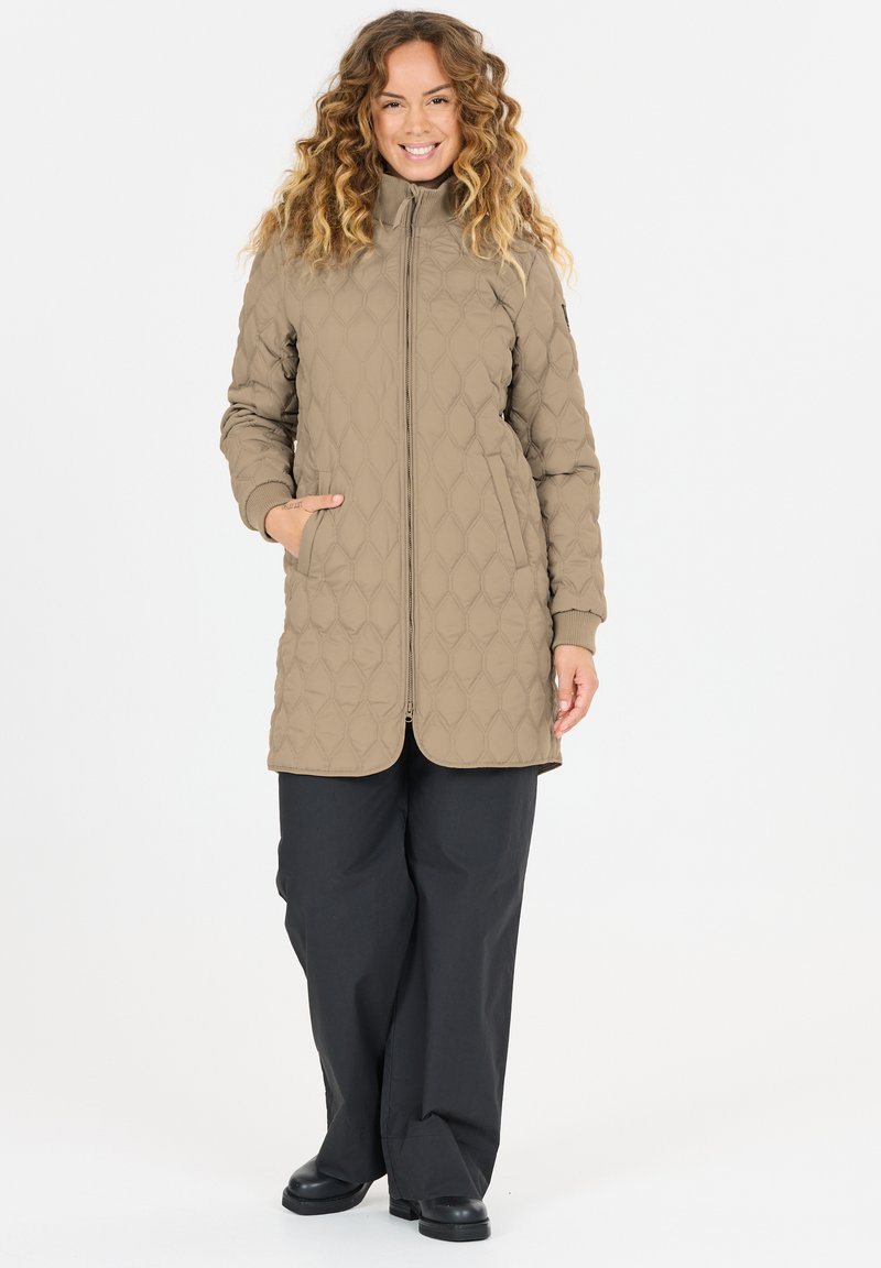 Vrouw met krullend haar die een doorgestikte beige lange jas draagt, zwarte wijde broekspijpen en zwarte schoenen, staand met één hand in de zak.