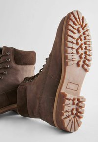 Botas de tobillo de piel marrón y ante con ojales de metal, suelas de goma gruesas que presentan un patrón de tracción robusto y soporte acolchado en el tobillo.
