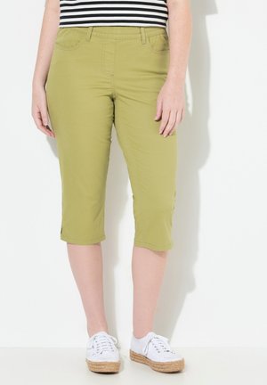 Olijfgroene capri-broek van rekbaar materiaal met zijzakken en een split aan de zoom, gecombineerd met witte sneakers en een zwart-wit gestreepte top.