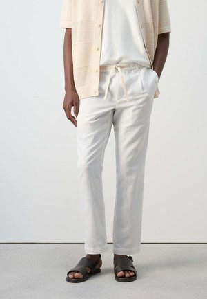 Uomo con maglietta a maniche corte in maglia beige chiaro, pantaloni bianchi con coulisse e sandali neri aperti, in piedi davanti a uno sfondo semplice.