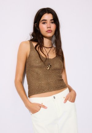 Jeune femme aux longs cheveux bruns porte un crop top en tricot marron sans manches, un pantalon blanc et un collier marron avec des pendentifs lune et étoile.