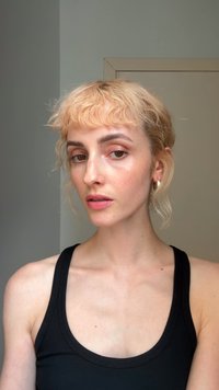 Portræt af en person med bølget, kort blont hår og lys hud, iført en sort tanktop. Trækkene inkluderer diskret makeup og hoops-øreringe.