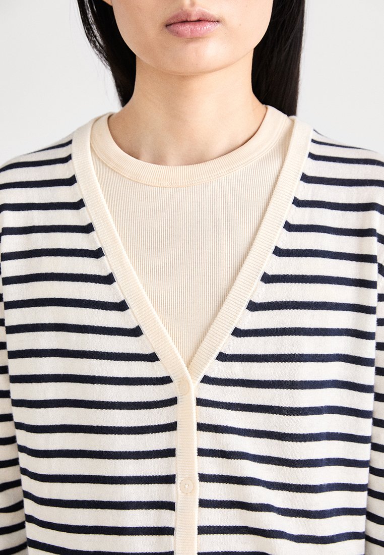 Marineblau- und cremegestreifter Cardigan mit V-Ausschnitt, Knopfleiste und leichtem Material, kombiniert mit einem gerippten, neutralen Oberteil.
