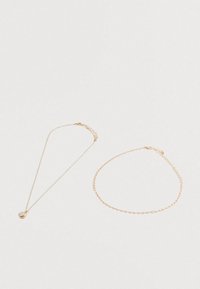 Dos delicadas cadenas de oro sobre una superficie blanca; una con un colgante circular de nácar, y la otra una sencilla pulsera de cadena.