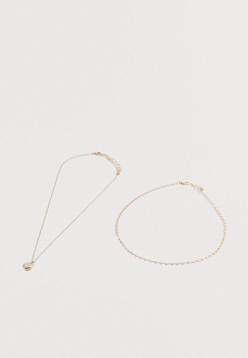 Dos delicadas cadenas de oro sobre una superficie blanca; una con un colgante circular de nácar, y la otra una sencilla pulsera de cadena.