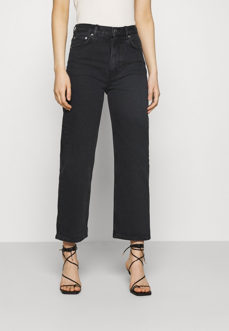 Personne portant un jean noir taille haute à jambes larges, associé à des sandales à talons noires à fines lanières et un haut blanc.