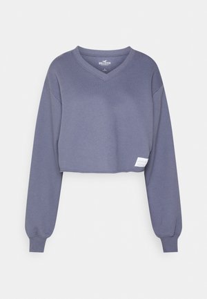 Bluza krótkiego kroju z długim rękawem i dekoltem w serek w stonowanym niebieskim kolorze, z prążkowanymi mankietami i małą białą prostokątną metką przy dole.