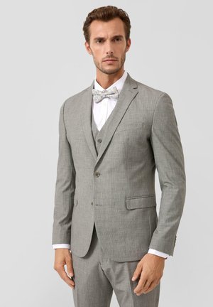 Man die een lichtgrijs driedelig pak draagt met een wit overhemd en een gestippelde strik, staand tegen een effen achtergrond.