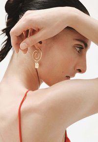 Mujer con el cabello oscuro recogido, usando un pendiente espiral de oro con un pequeño charm rectangular, vestida con una blusa de tirantes rojos.